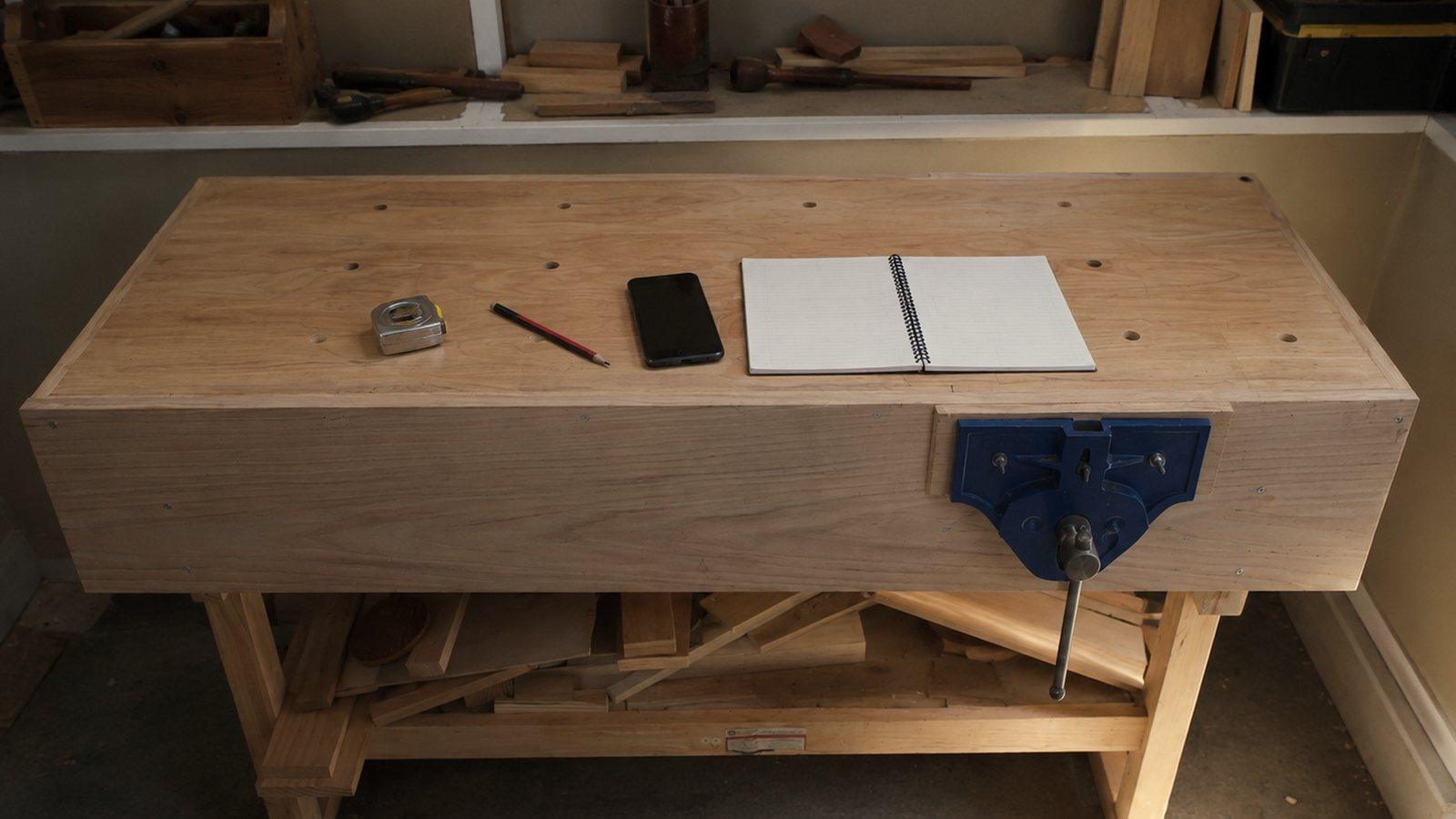 A timber workbench in morning light with a tape measure, a pencil, a phone and a notepad on it