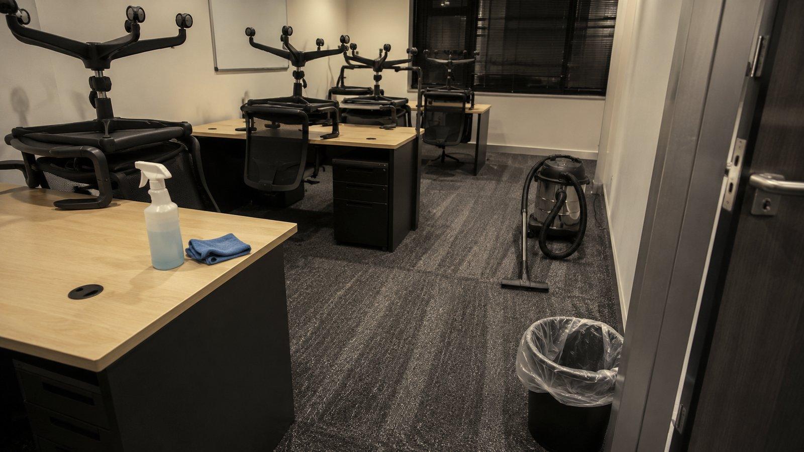 An office with the chairs up on the desks and a vacuum on the carpet, mid clean