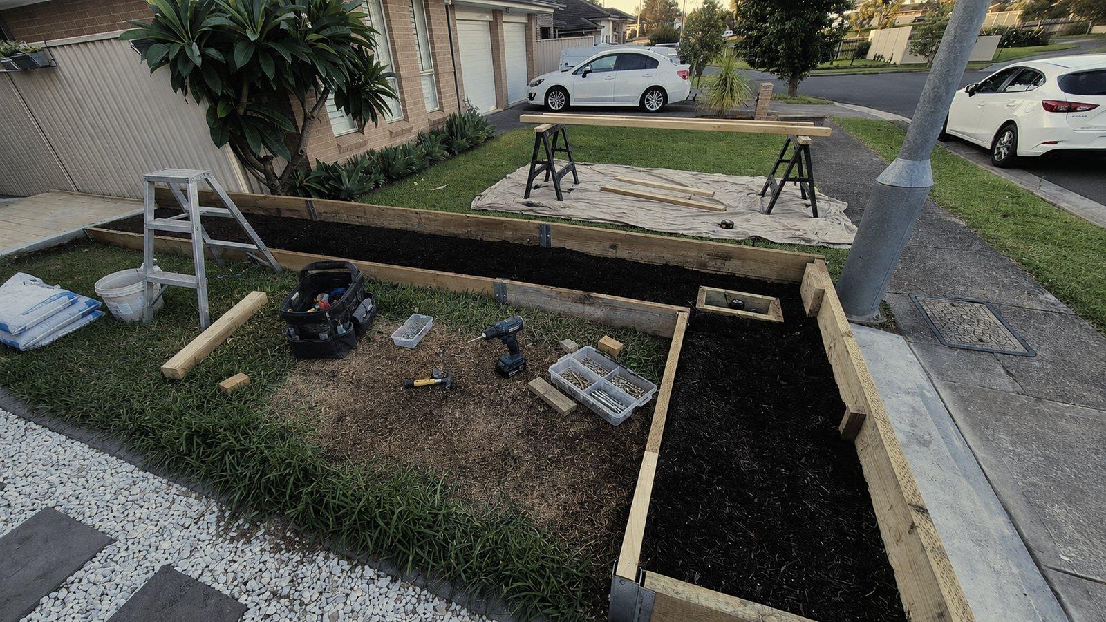 Timber garden bed edging part way through a build along a front lawn, sawhorses and a tool bag on the grass